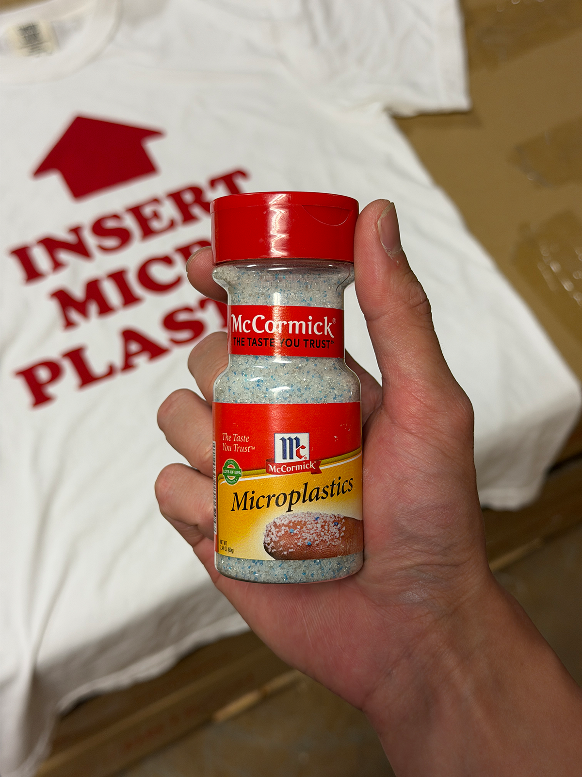 Hand holding a bottle of McCormick Microplastics seasoning with a shirt in the background. Shirt says INSERT MICRO PLASTICS