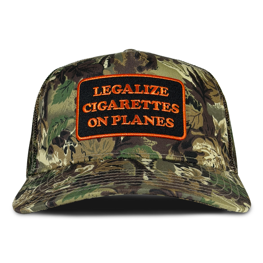 A camouflage trucker-style cap with a front patch reading 'LEGALIZE CIGARETTES ON PLANES' in orange text on a black background, shown against a plain white background. Hat by Crying in the Club Shirts