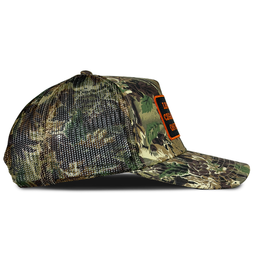 A camouflage trucker cap with a mesh back and a small orange patch on the side, shown against a plain white background. Hat by Crying in the Club Shirts