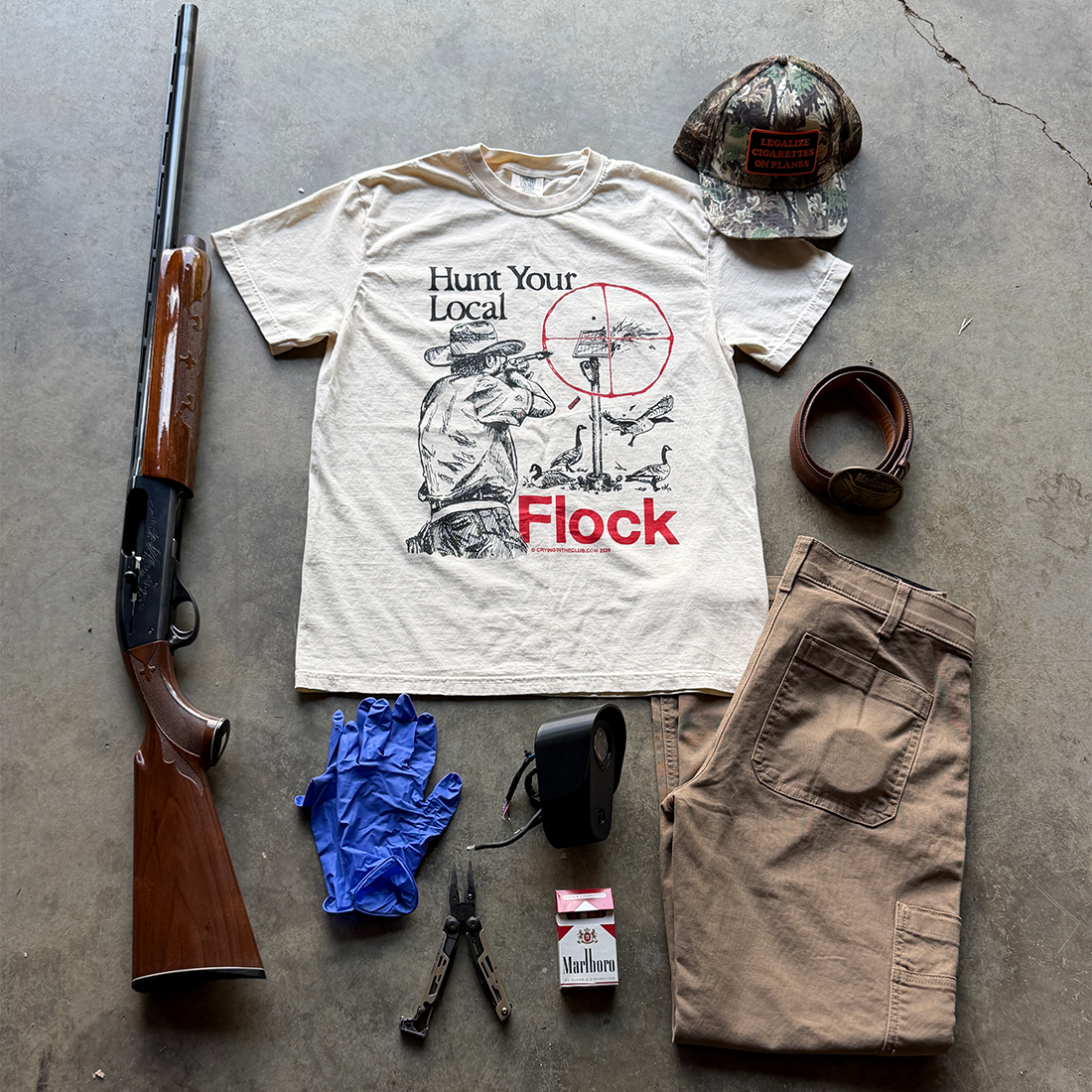 A flat lay of a vintage-style hunting t-shirt with a 'Hunt Your Local Flock' graphic, styled with a shotgun, camouflage cap, tan pants, blue gloves, pliers, a flashlight, and a pack of cigarettes on a concrete floor. Crying in the Club T Shirts 