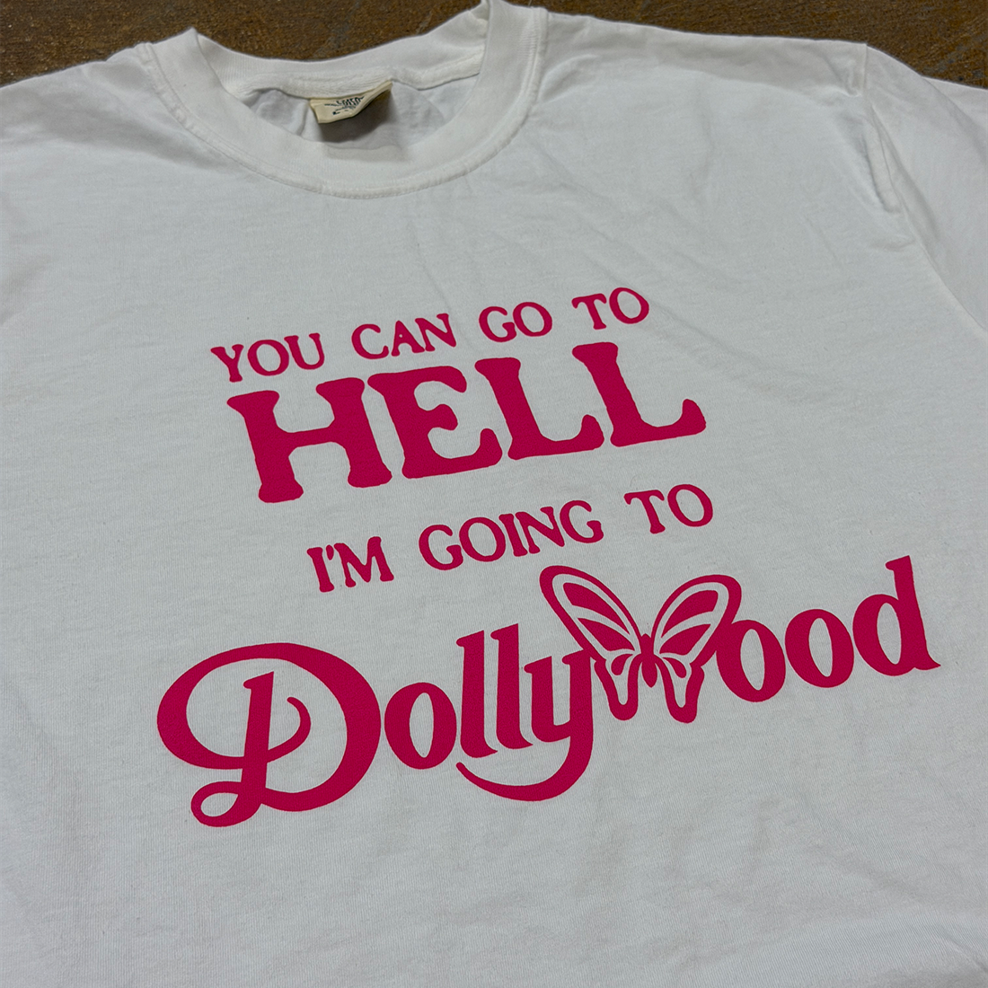 A white short-sleeve t-shirt with a bold pink graphic text that reads 'YOU CAN GO TO HELL I'M GOING TO Dollywood' with a butterfly graphic, laid flat on a brown surface.