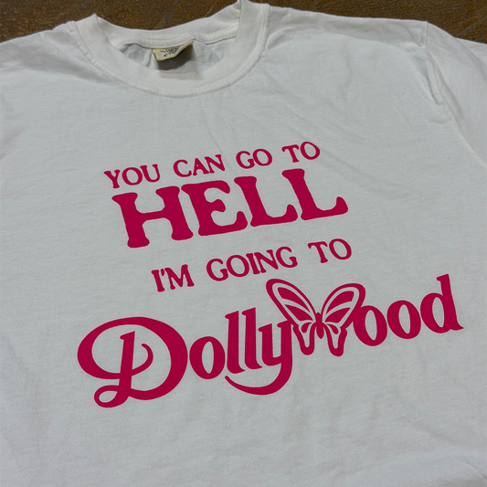 A white short-sleeve t-shirt with a bold pink graphic text that reads 'YOU CAN GO TO HELL I'M GOING TO Dollywood' with a butterfly graphic, laid flat on a brown surface.