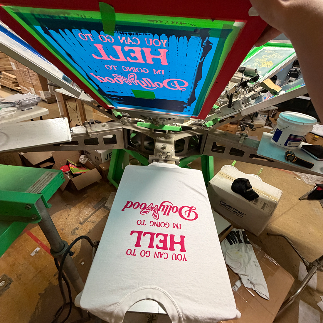 A white t-shirt is being printed on a screen in a workshop setting. The screen shows a bright blue background with pink and white text that reads 'YOU CAN GO TO HELL I'M GOING TO Dollywood' (mirrored).